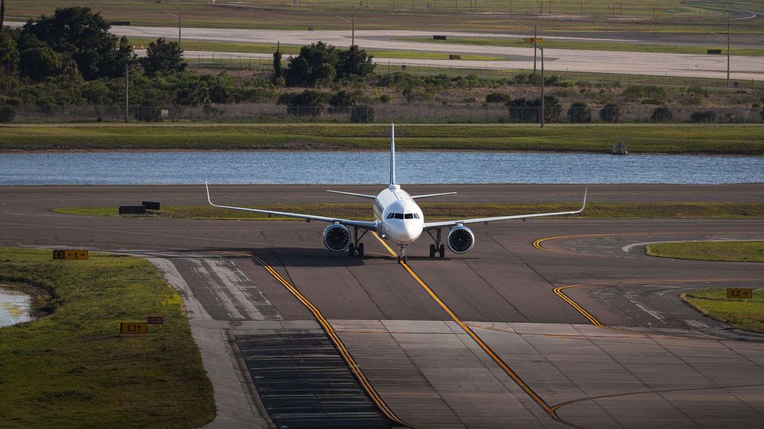 Commercial airplane taxiing on runway at airport with green landscape and pond in background.