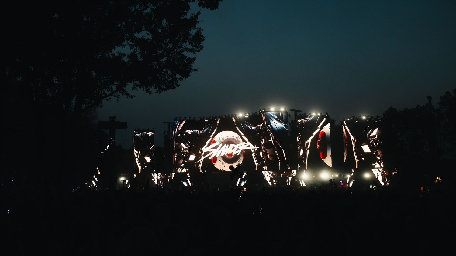 Silhouetted crowd with raised hands at an outdoor concert against a dark night sky with stage lights glowing in background.
