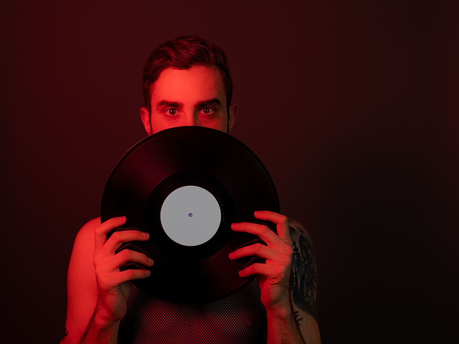Man holding vinyl record in front of face under dramatic red lighting, with visible tattoo on hand.