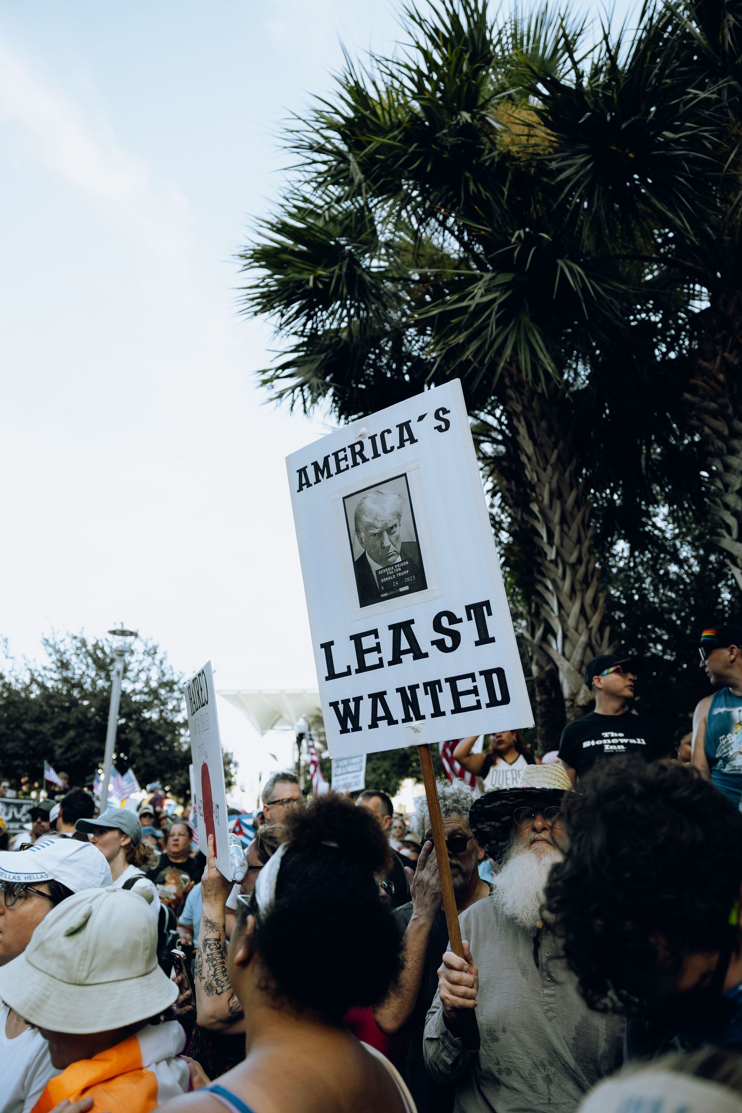 Protest sign reading 'America's Least Wanted' held up among crowd with palm trees in background.