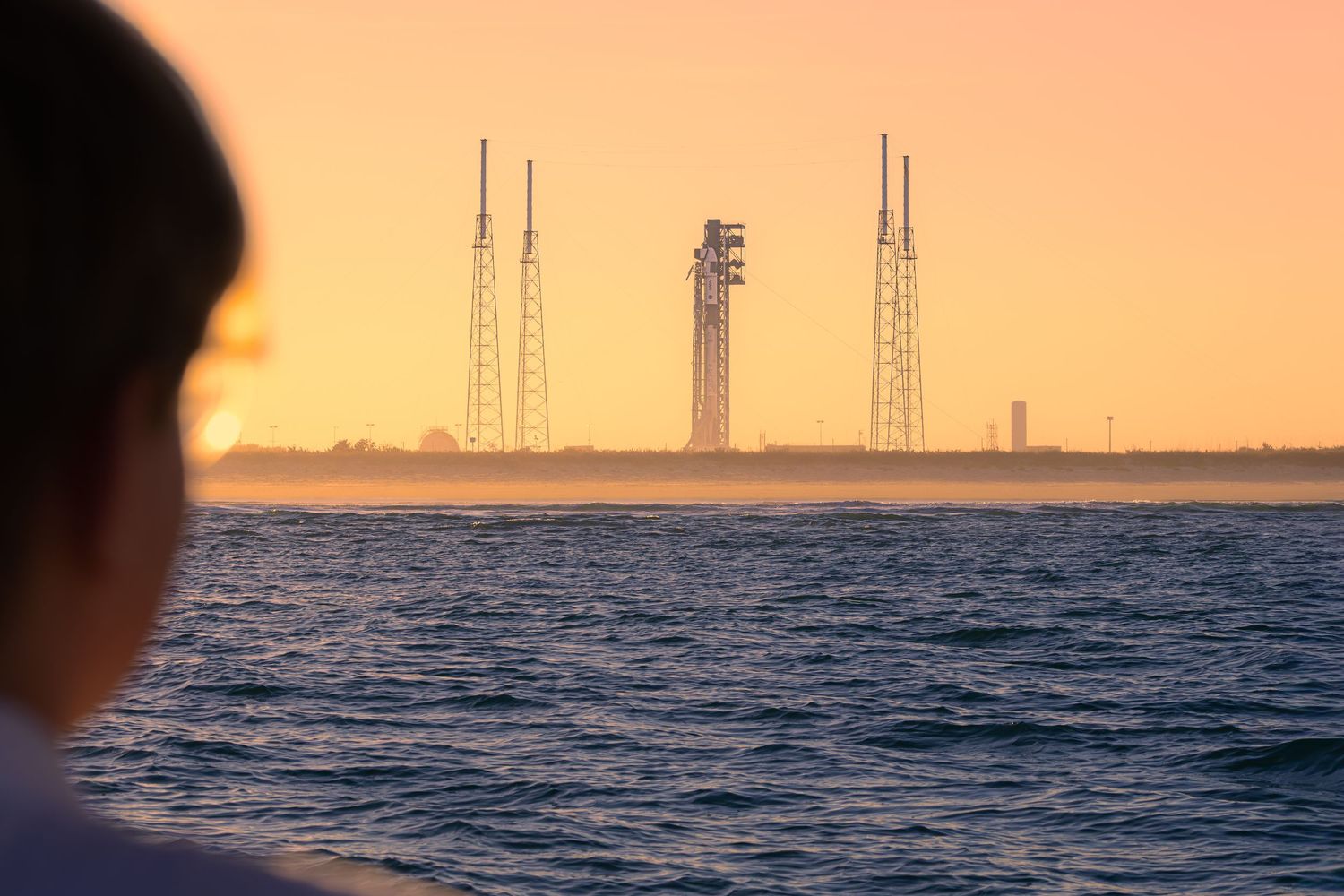 Rocket on launch pad at sunset viewed from water with warm orange sky and a person in foreground.