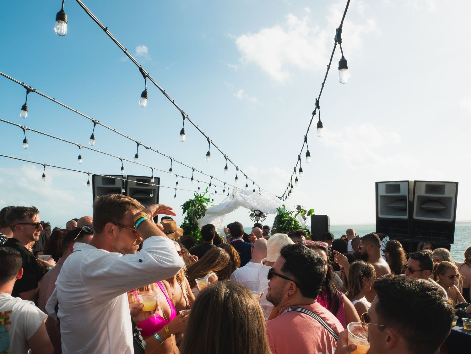 Large outdoor party crowd dancing under string lights at a daytime rooftop event with large speakers.