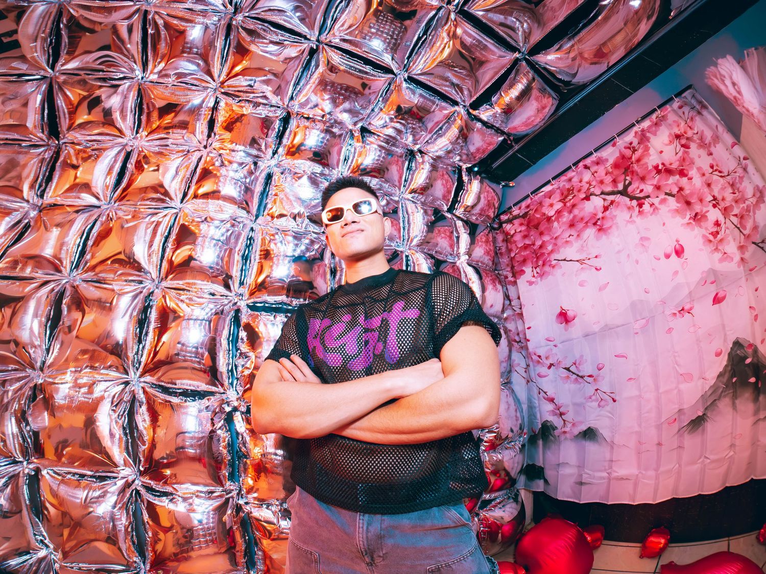 Man with crossed arms poses confidently in front of silver balloon wall and floral backdrop in nightclub.