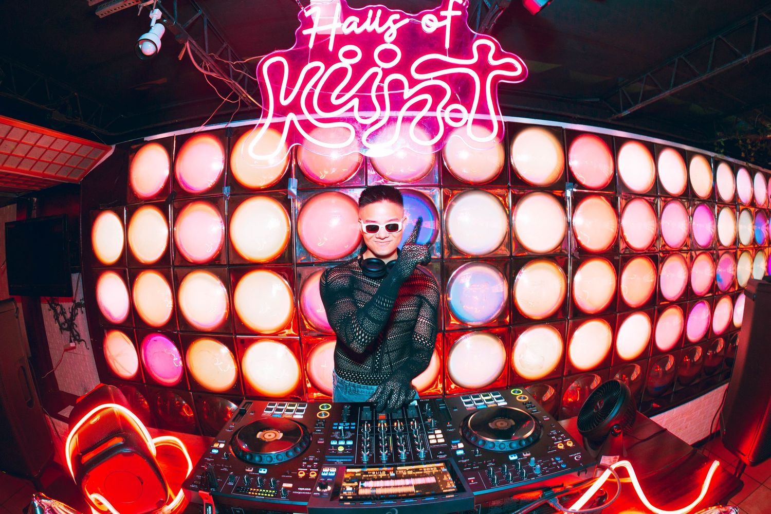 A DJ performing behind decks at Hall of Kunst club, surrounded by large round orange stage lights and neon signs.