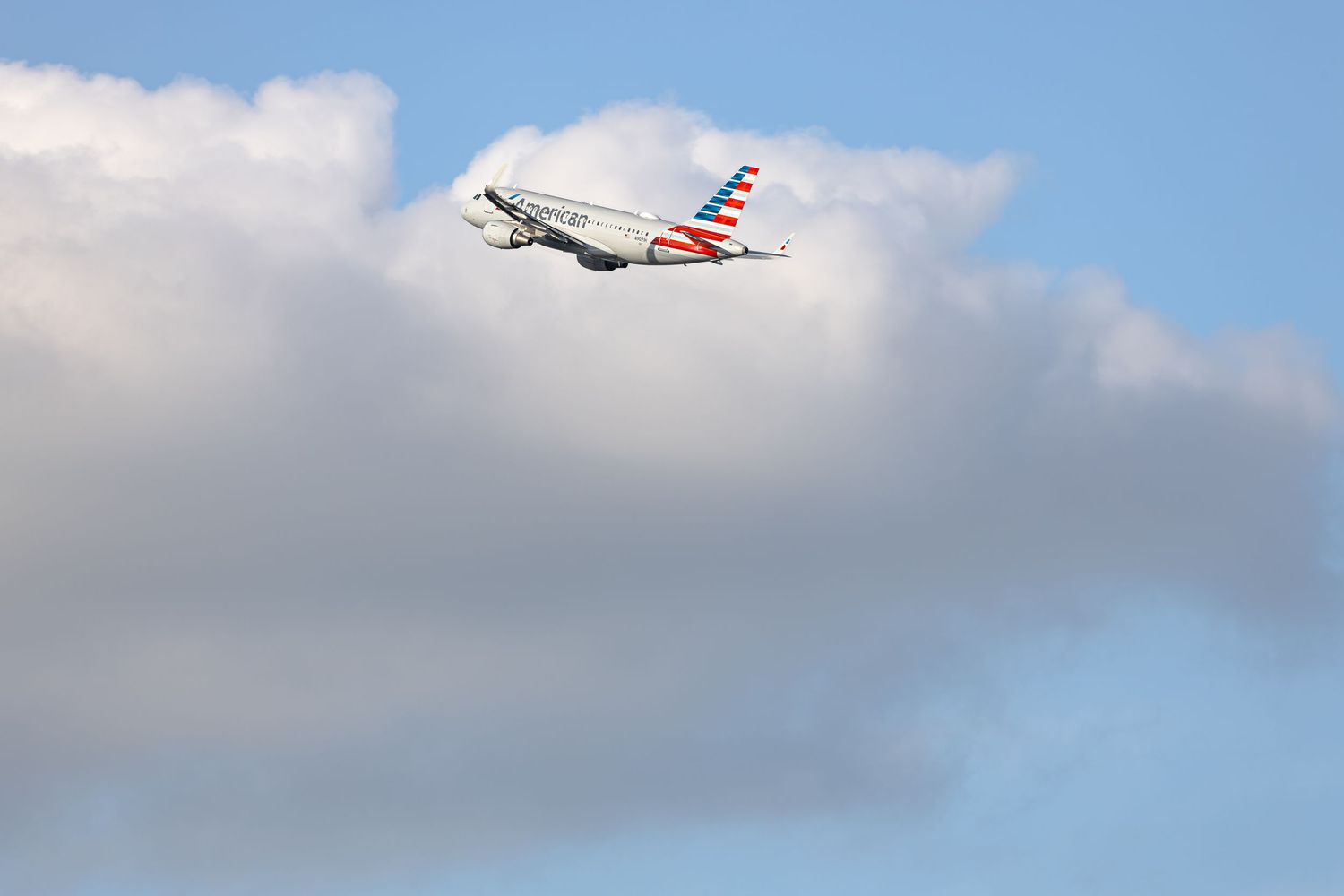American Airlines passenger jet climbing through blue sky with white clouds after takeoff.