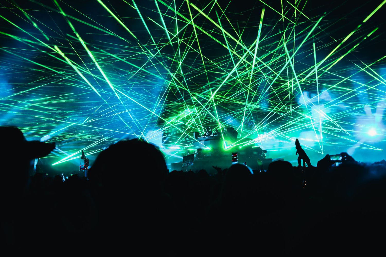 Vibrant green and blue laser beams illuminate a concert venue with silhouetted crowd against a dark background.