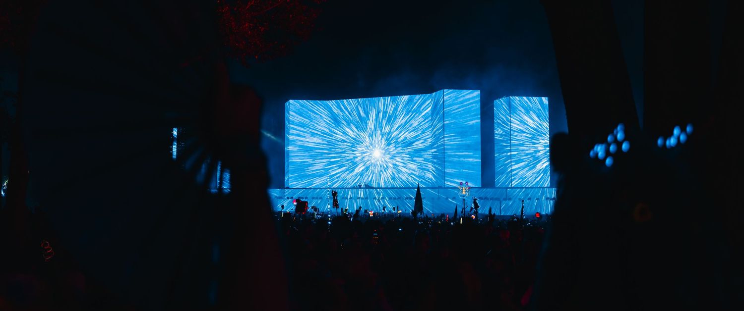 A dark nighttime scene shows glowing blue lights reflecting off water with silhouettes of trees in the background.