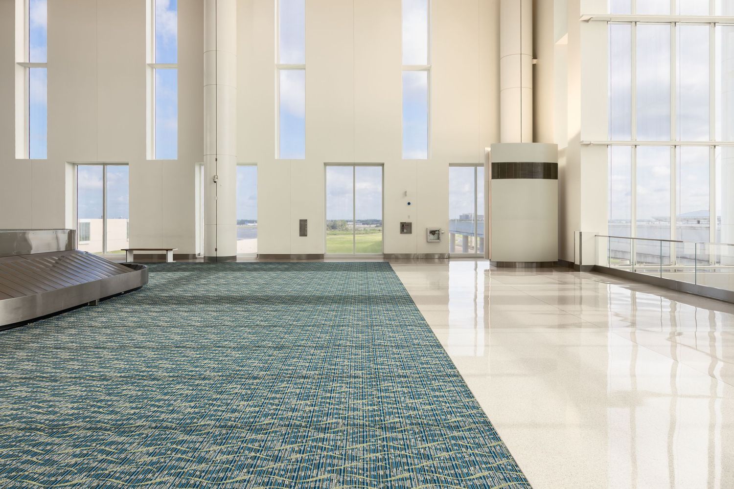 Spacious airport terminal with teal patterned carpet, white tile floors, large windows, and high ceilings.