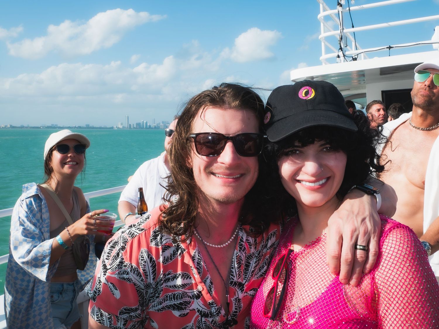 People enjoying a sunny day on a boat deck with turquoise waters and city skyline visible in the background.