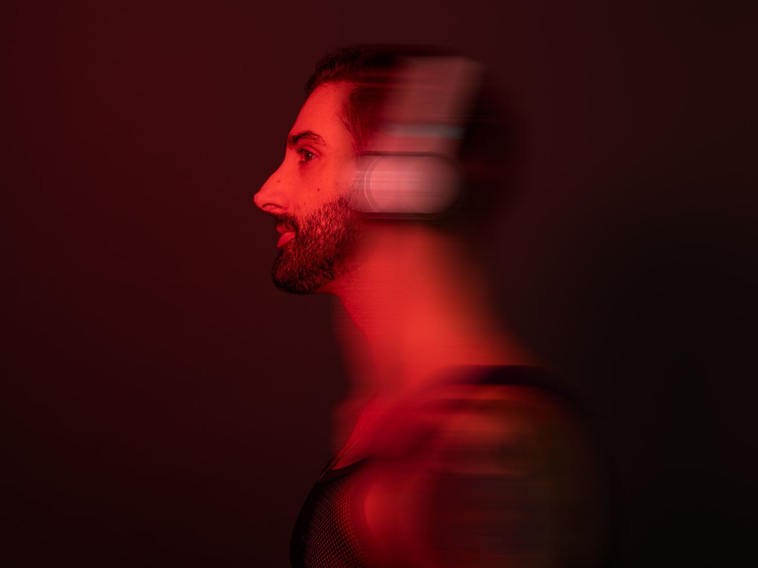 A bearded man in profile view bathed in dramatic red light with a motion blur effect on his face.