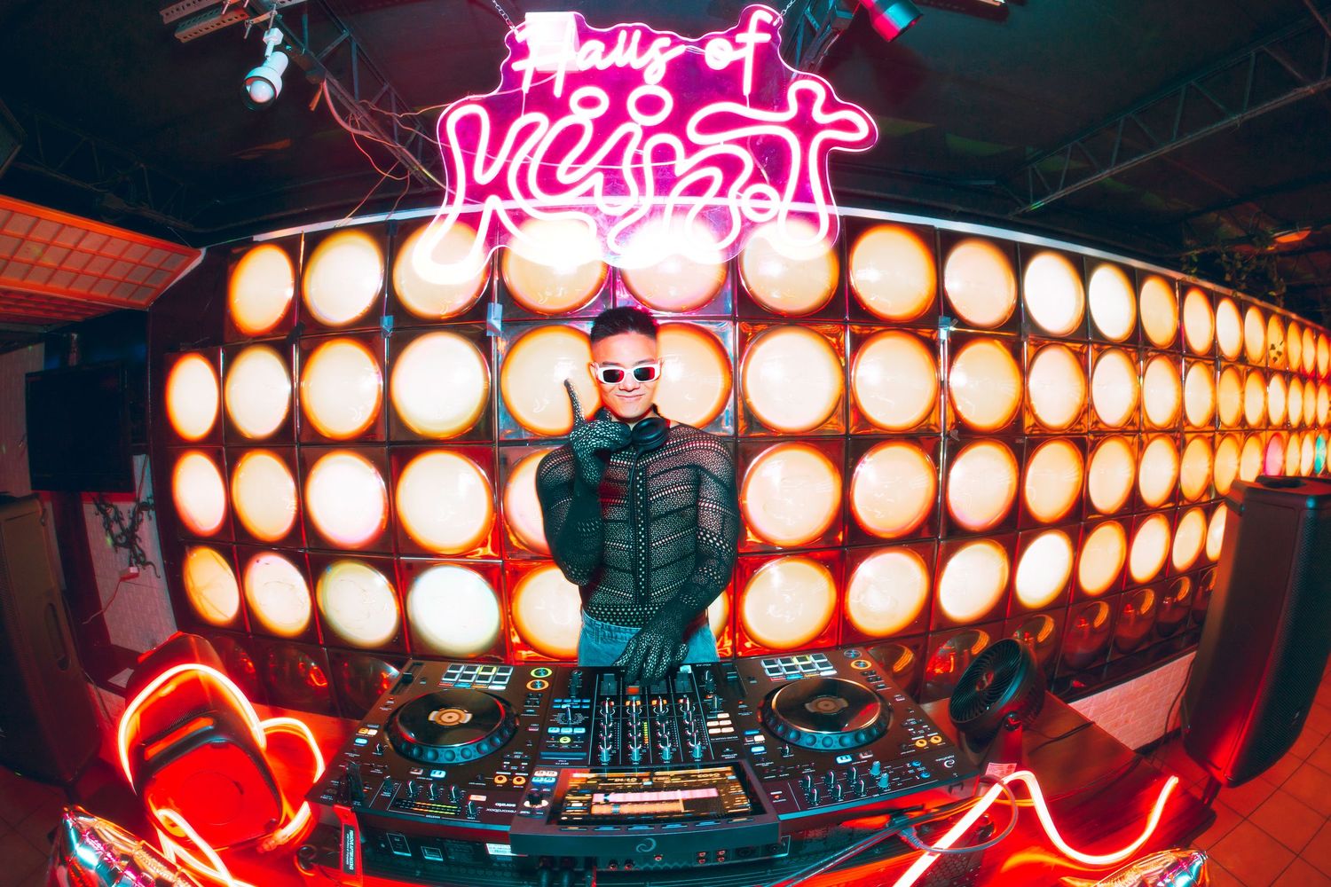 DJ performing behind decks with neon lights and illuminated wall backdrop in vibrant nightclub setting.