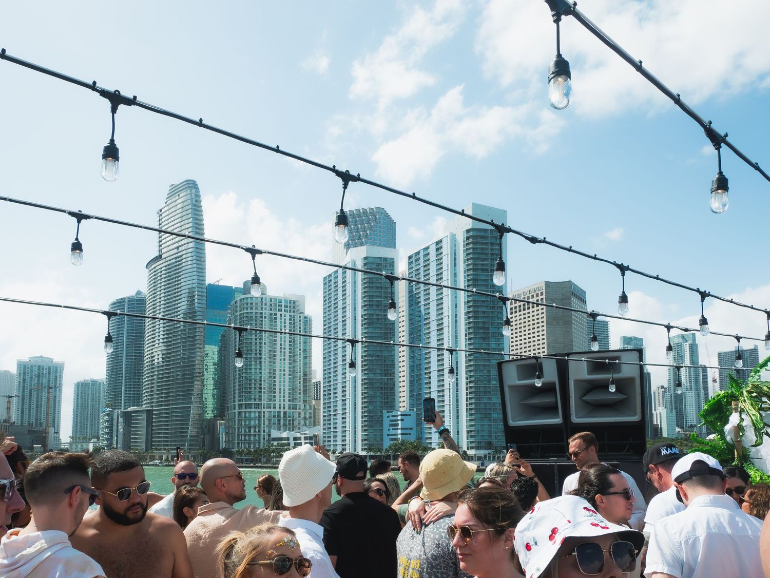 Rooftop party crowd enjoys city views with string lights and Miami downtown skyscrapers in the background.