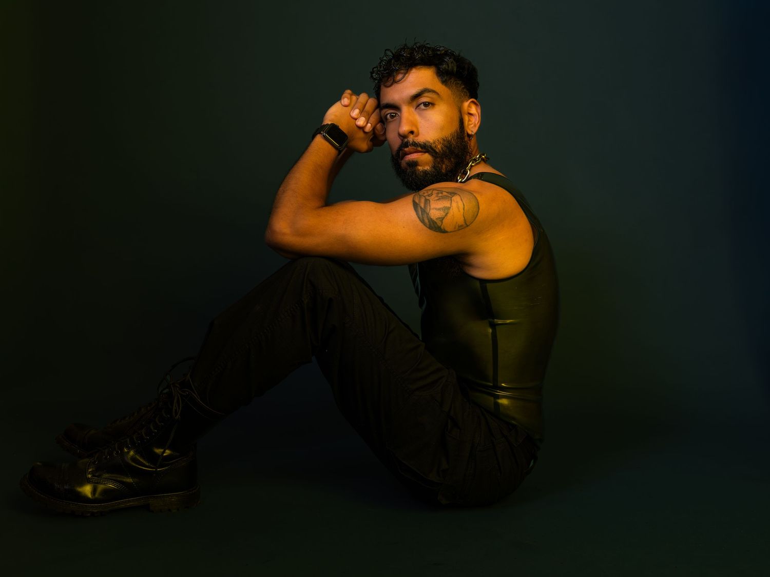 Tattooed man in black sleeveless outfit sits on floor, posing with hand near face against dark teal background.