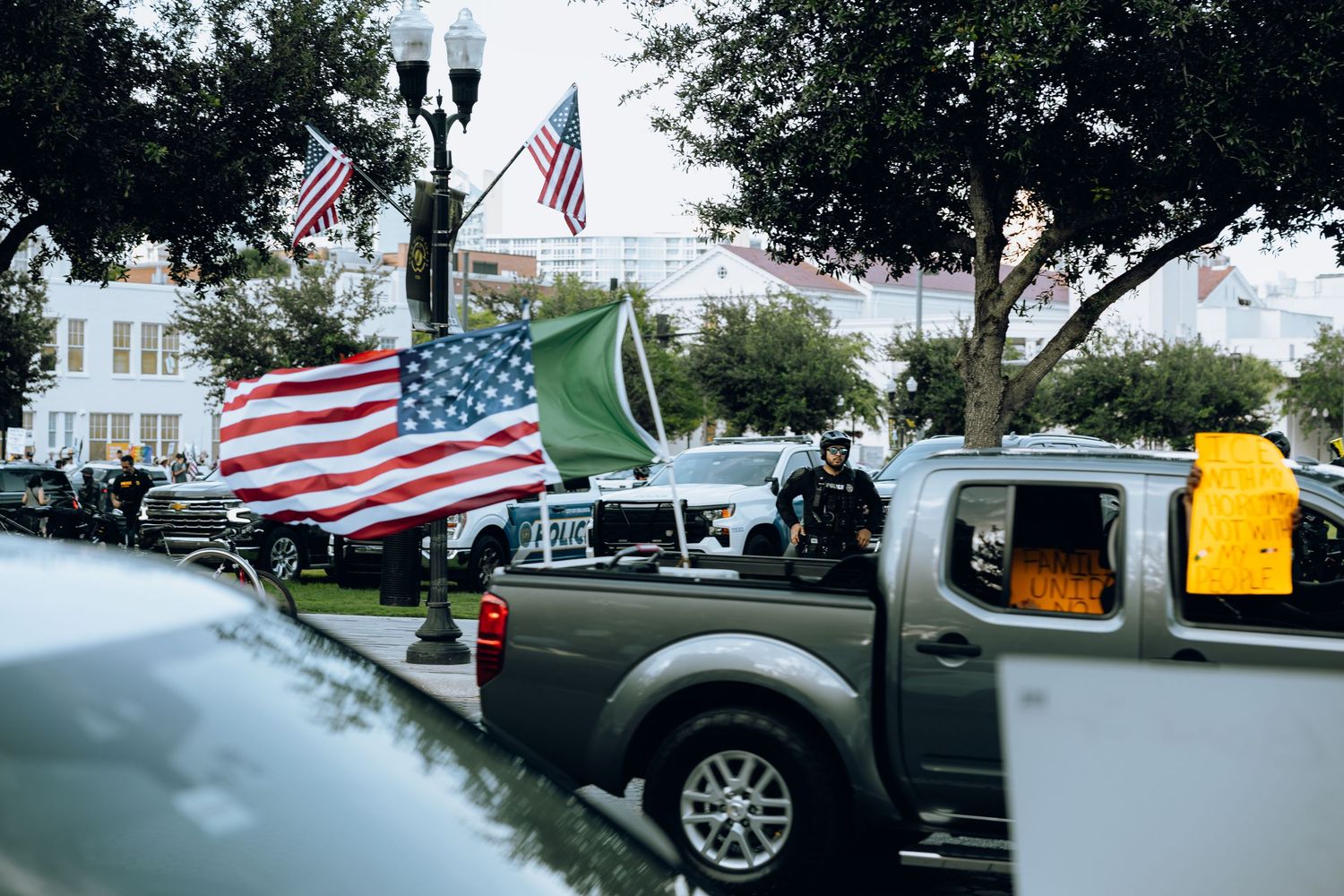American flags mounted on pickup trucks parked along a street with trees and buildings visible.