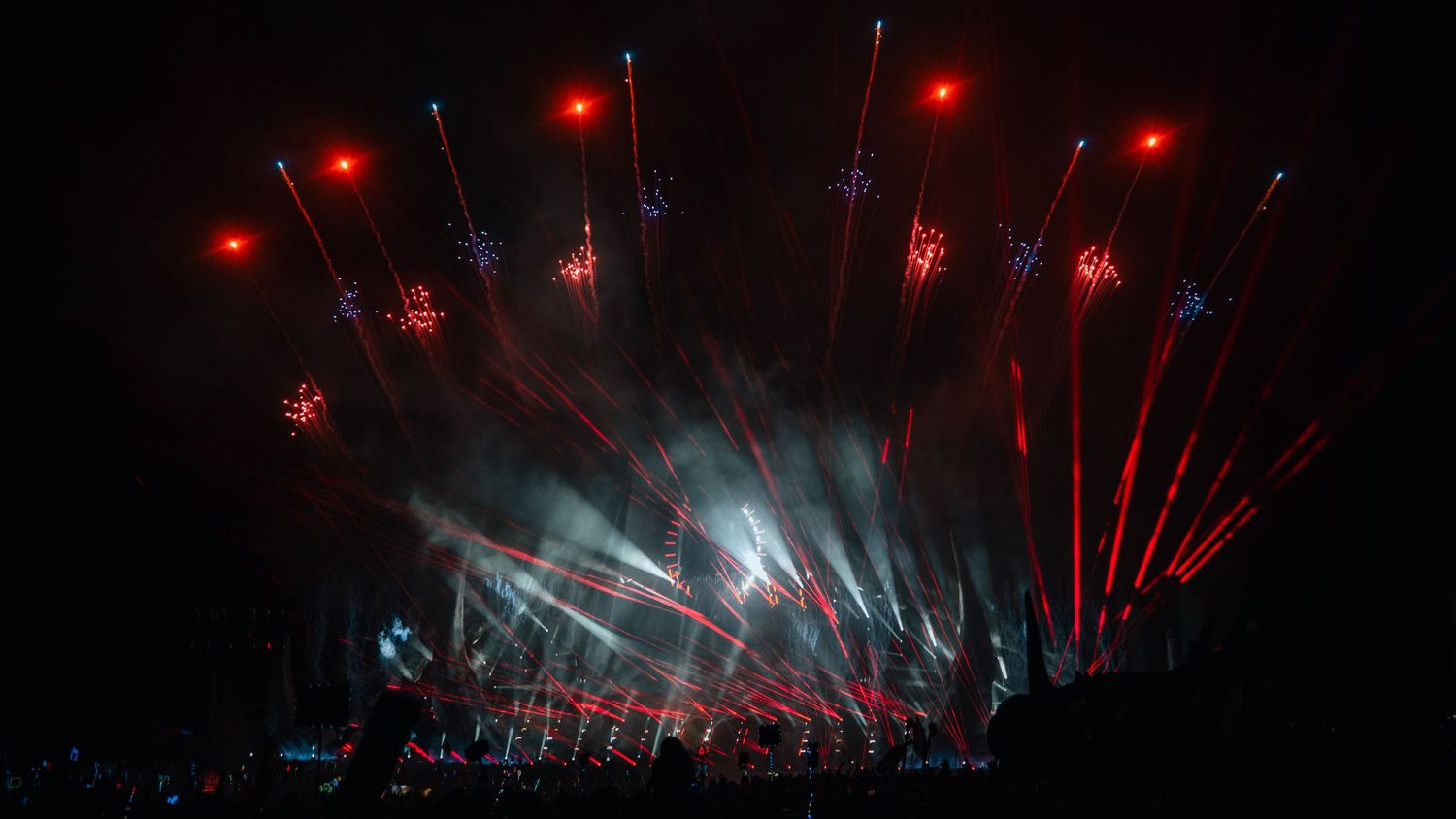 Red laser beams illuminate a dark concert stage with silhouetted performers and atmospheric lighting effects.