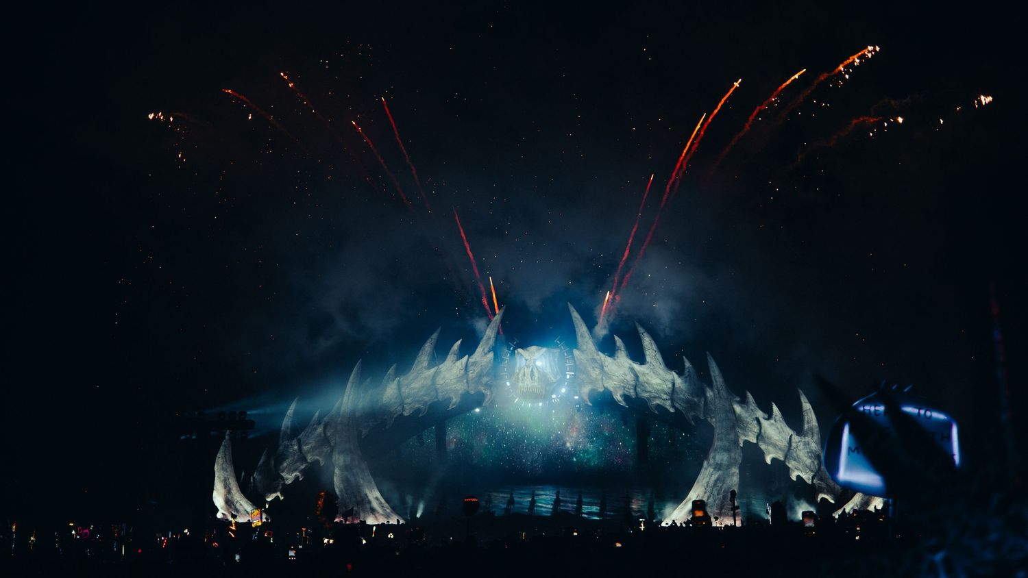 Dramatic concert stage with blue and white lighting effects and red firework trails illuminating the dark arena.