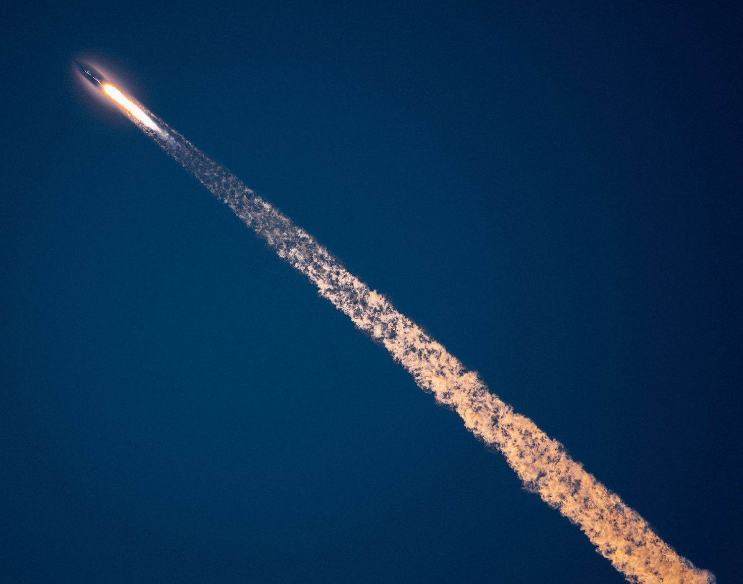 A rocket launches into a deep blue sky leaving a long trail of white and golden exhaust smoke behind it.