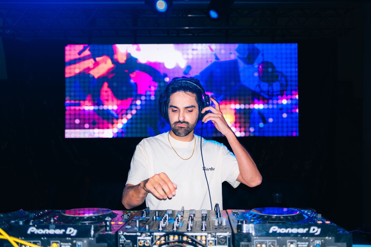 A DJ performs behind turntables at a nightclub with colorful blue and pink lighting effects in the background.