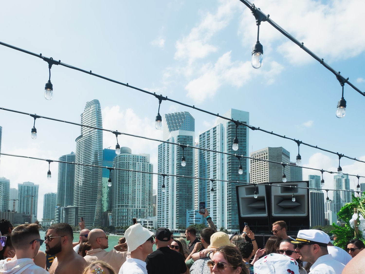 Crowded outdoor rooftop party with string lights and Miami city skyline featuring modern skyscrapers in background.