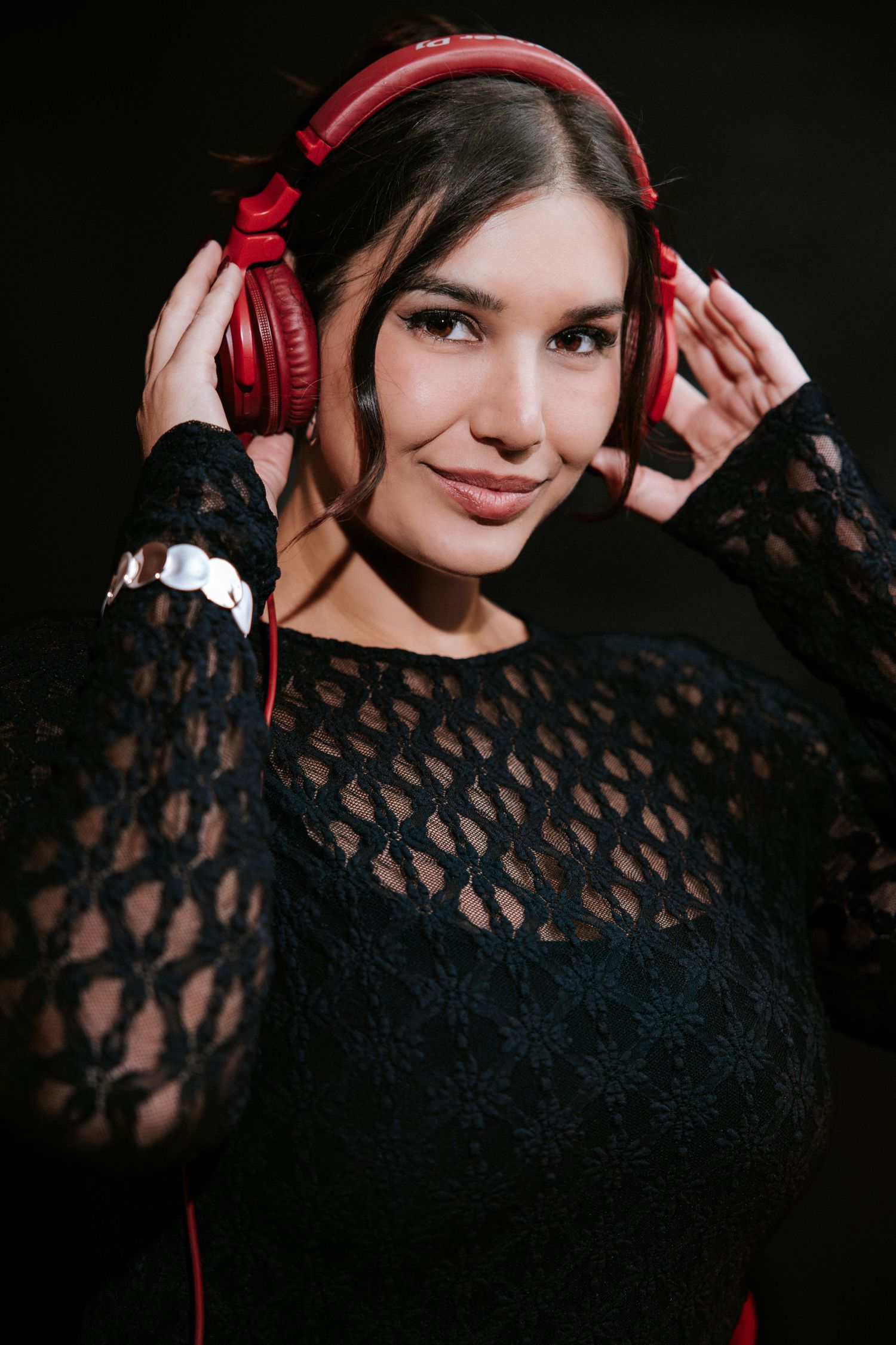 Young woman wearing red headphones and black lace top, smiling at camera against dark background.