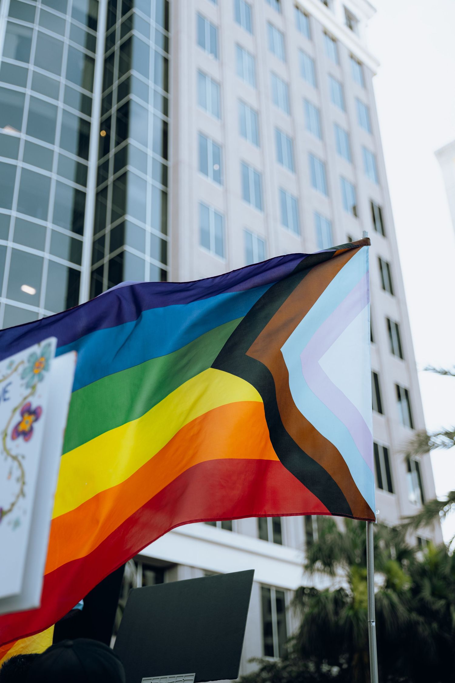 Progress pride flag waving in front of a modern glass office building on a sunny day.