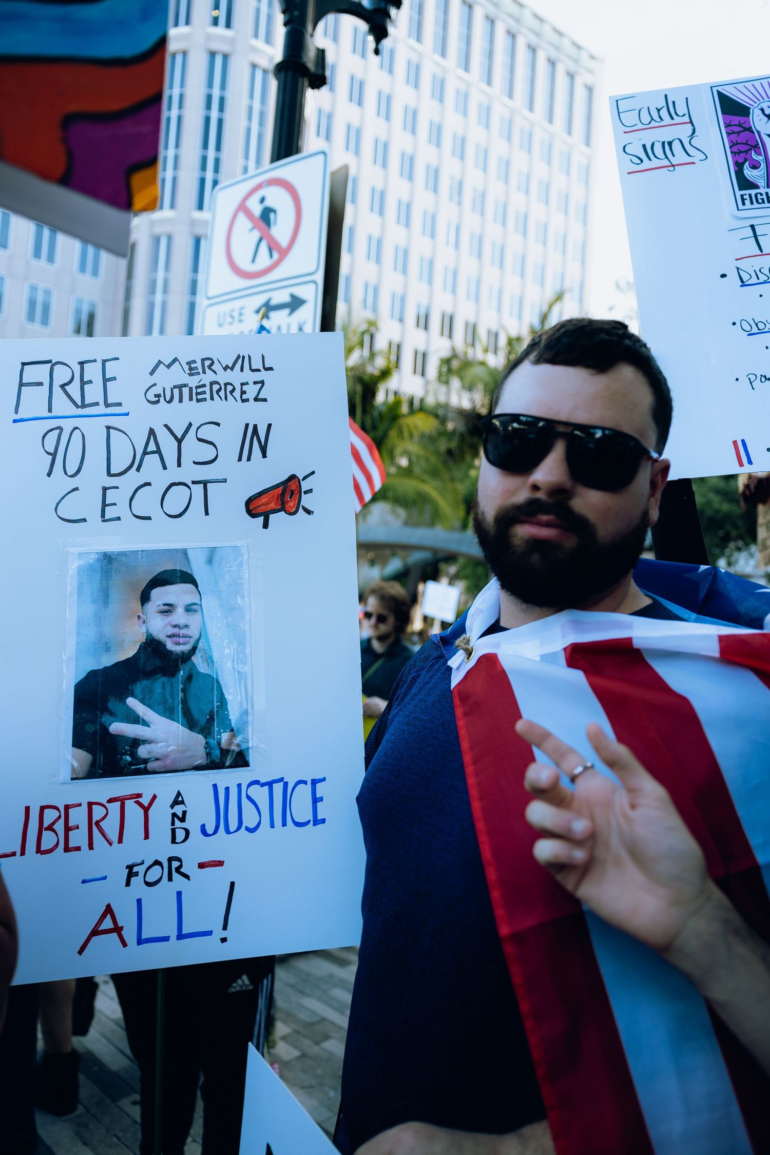 Protester wrapped in American flag stands next to sign advocating for liberty and justice.