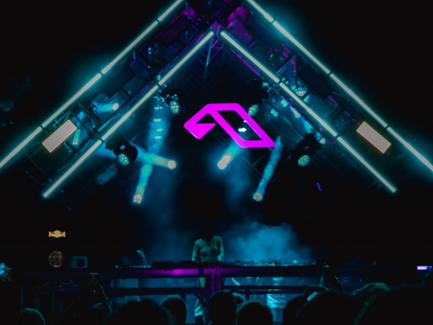Neon pink geometric logo glows above a dark concert stage with blue lighting and electronic music equipment.