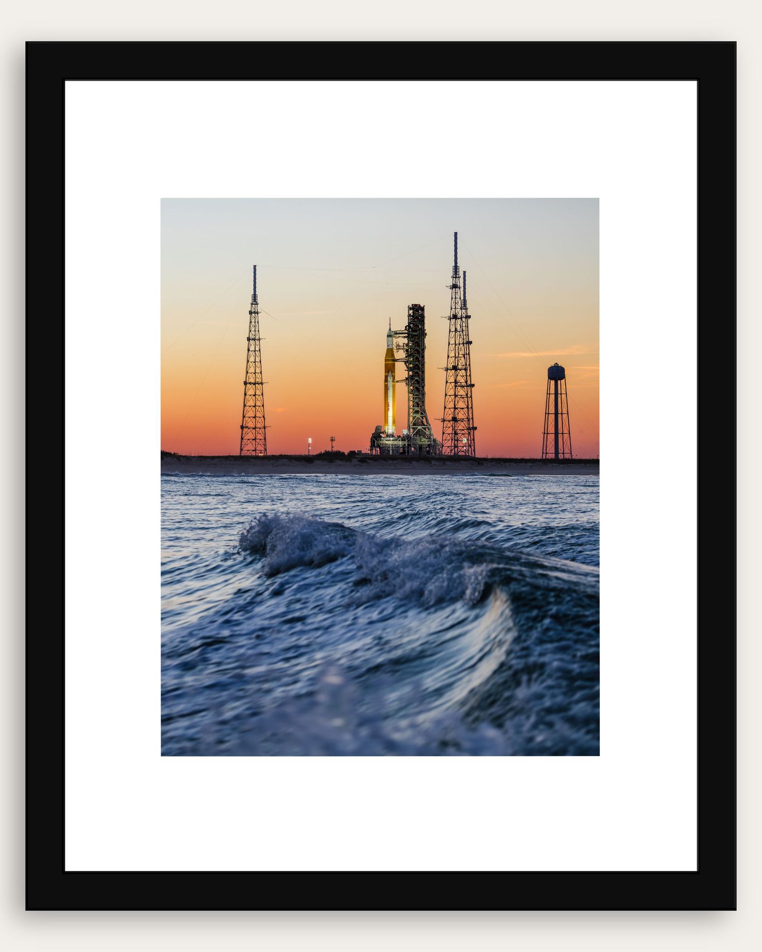 Framed photo of a NASA rocket on launch pad at sunset with ocean waves in the foreground at Kennedy Space Center.