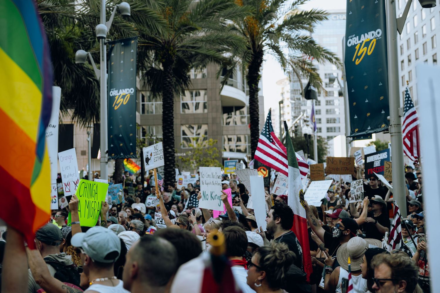 Large crowd of protesters gather in downtown with rainbow pride flag and American flags visible among palm trees.
