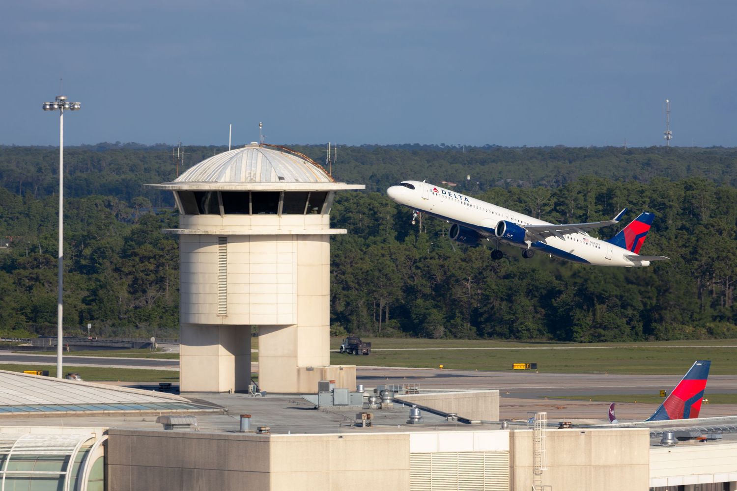 Delta Air Lines jet takes off near airport control tower with tree line in background on clear day.