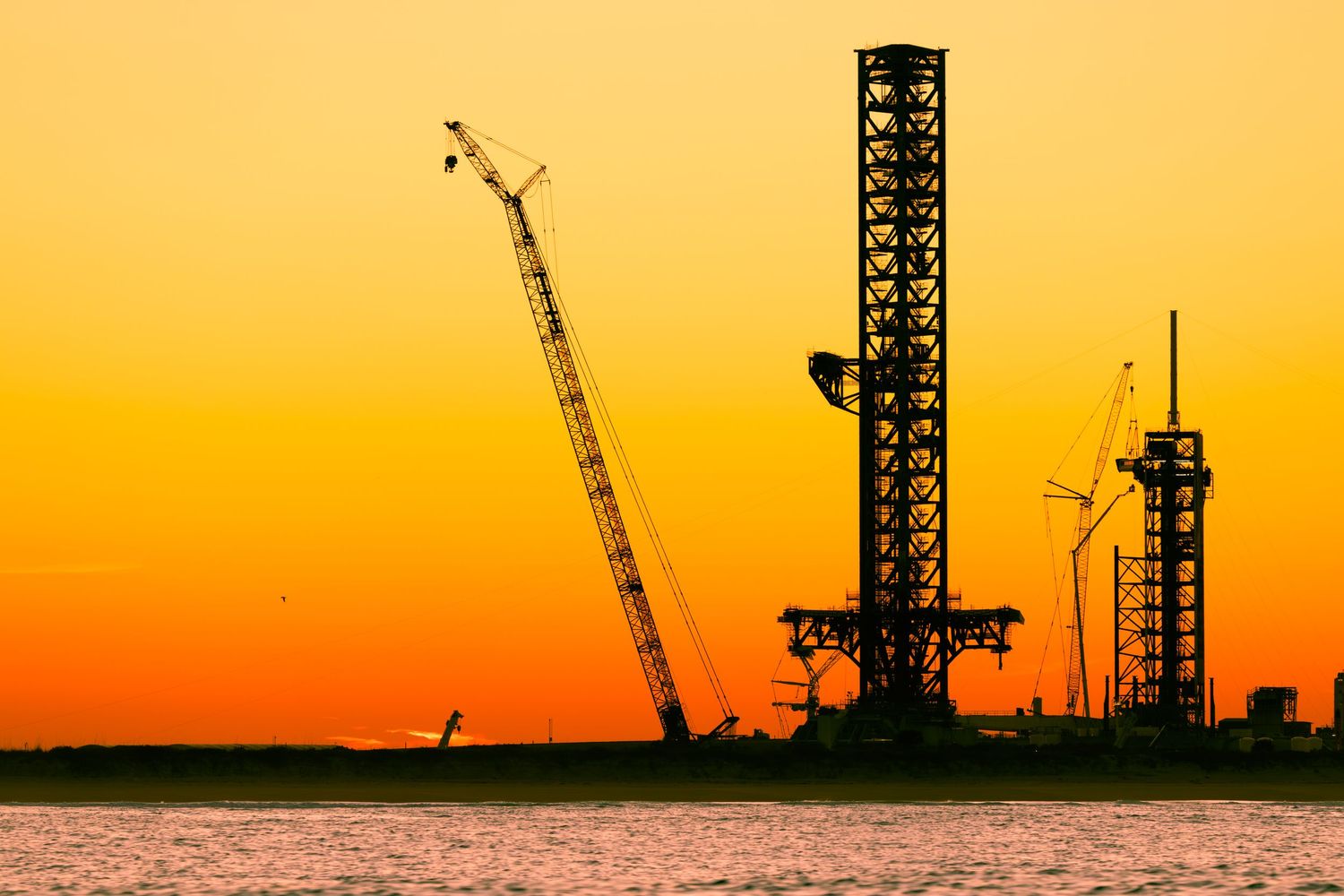 Silhouetted oil rig and cranes against a vivid orange sunset sky reflected over calm water.