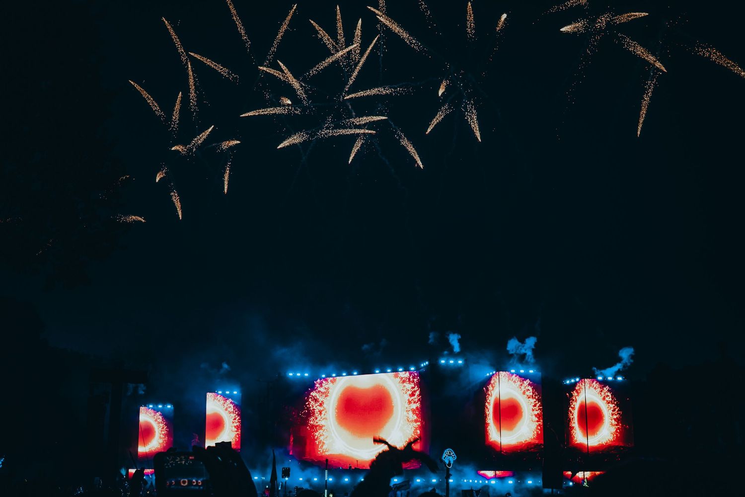 Colorful stage lighting display with red illuminated screens and firework effects against dark night sky.