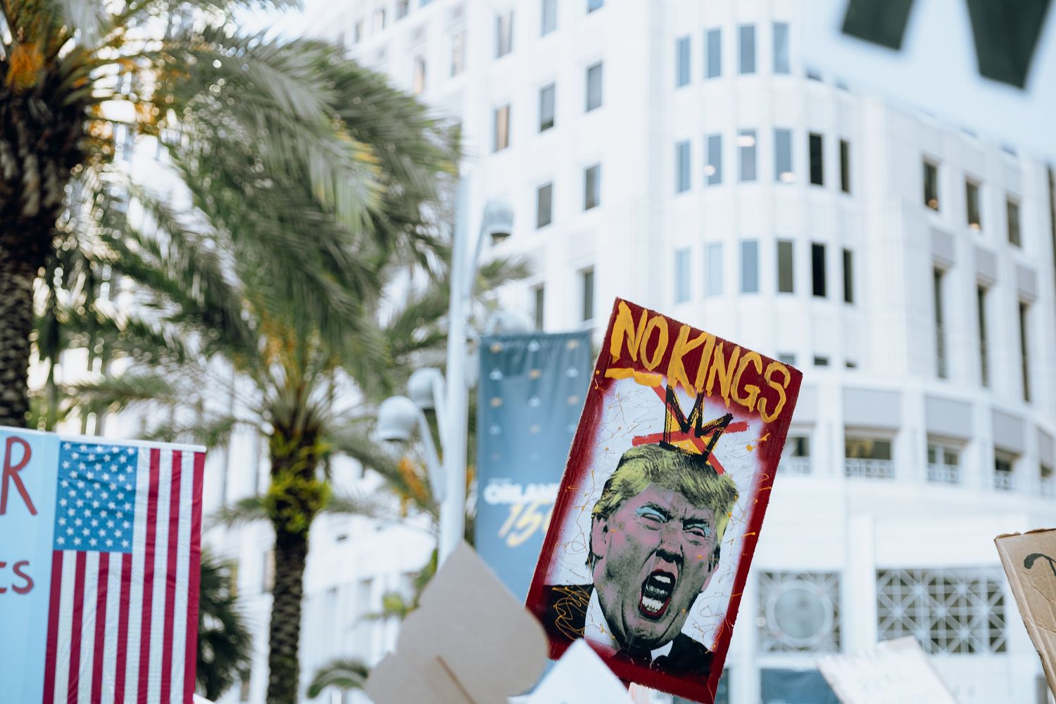 Protest signs and American flag against white office building with palm trees in urban setting.