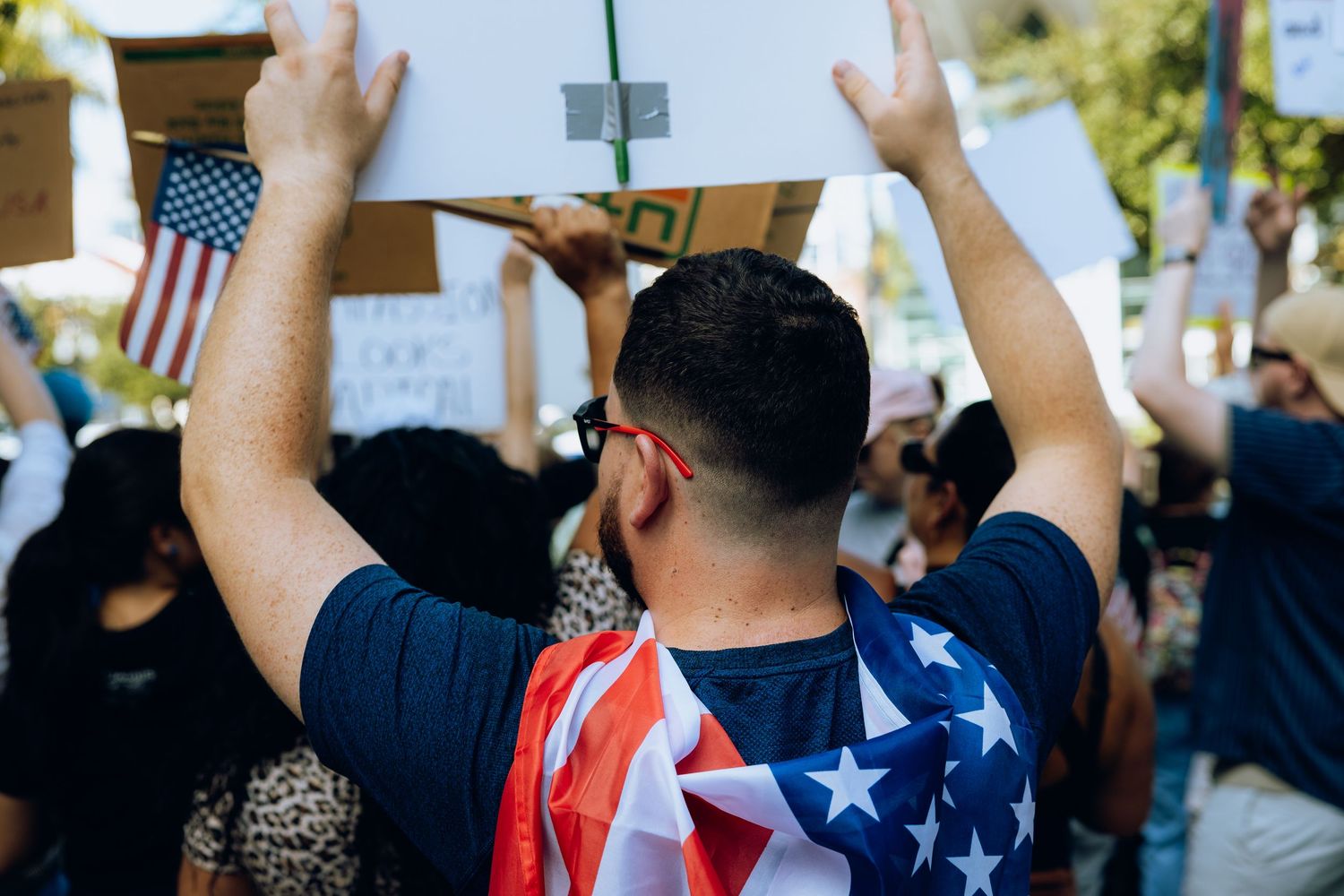Person in an American flag cape raises hands during a street protest surrounded by other demonstrators.