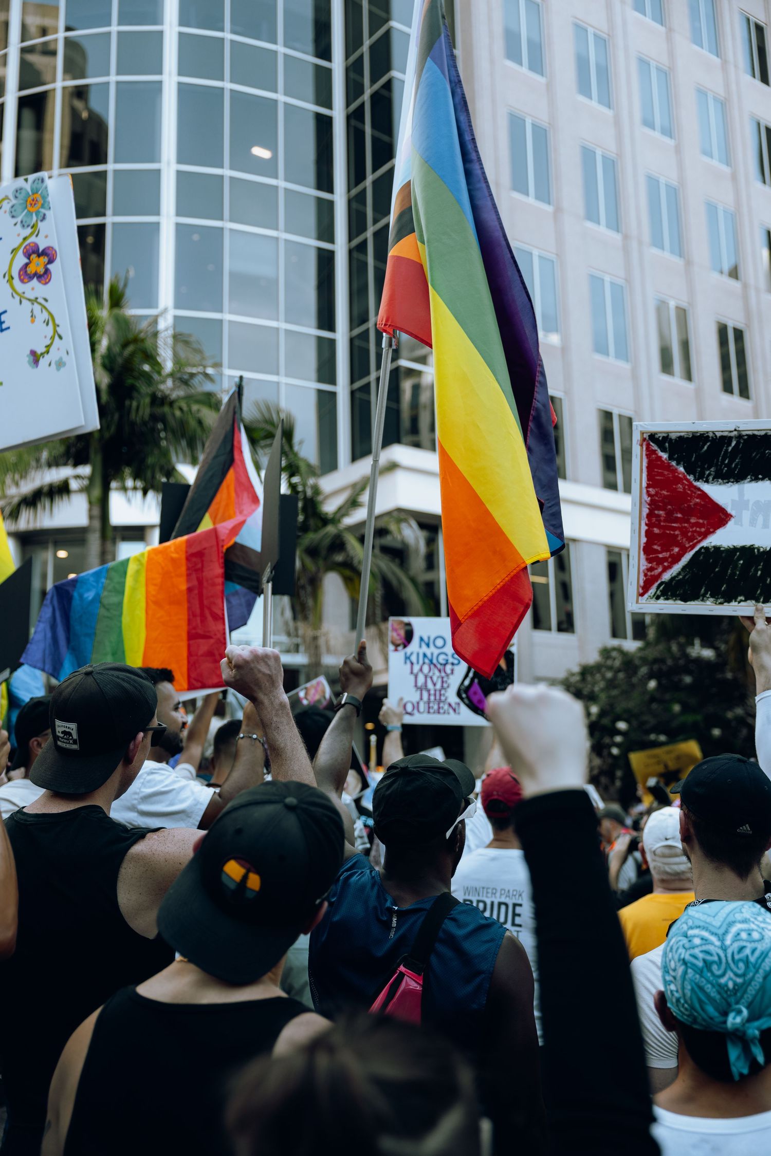 LGBTQ+ pride flags wave among a crowd of protesters gathered in front of an urban building.