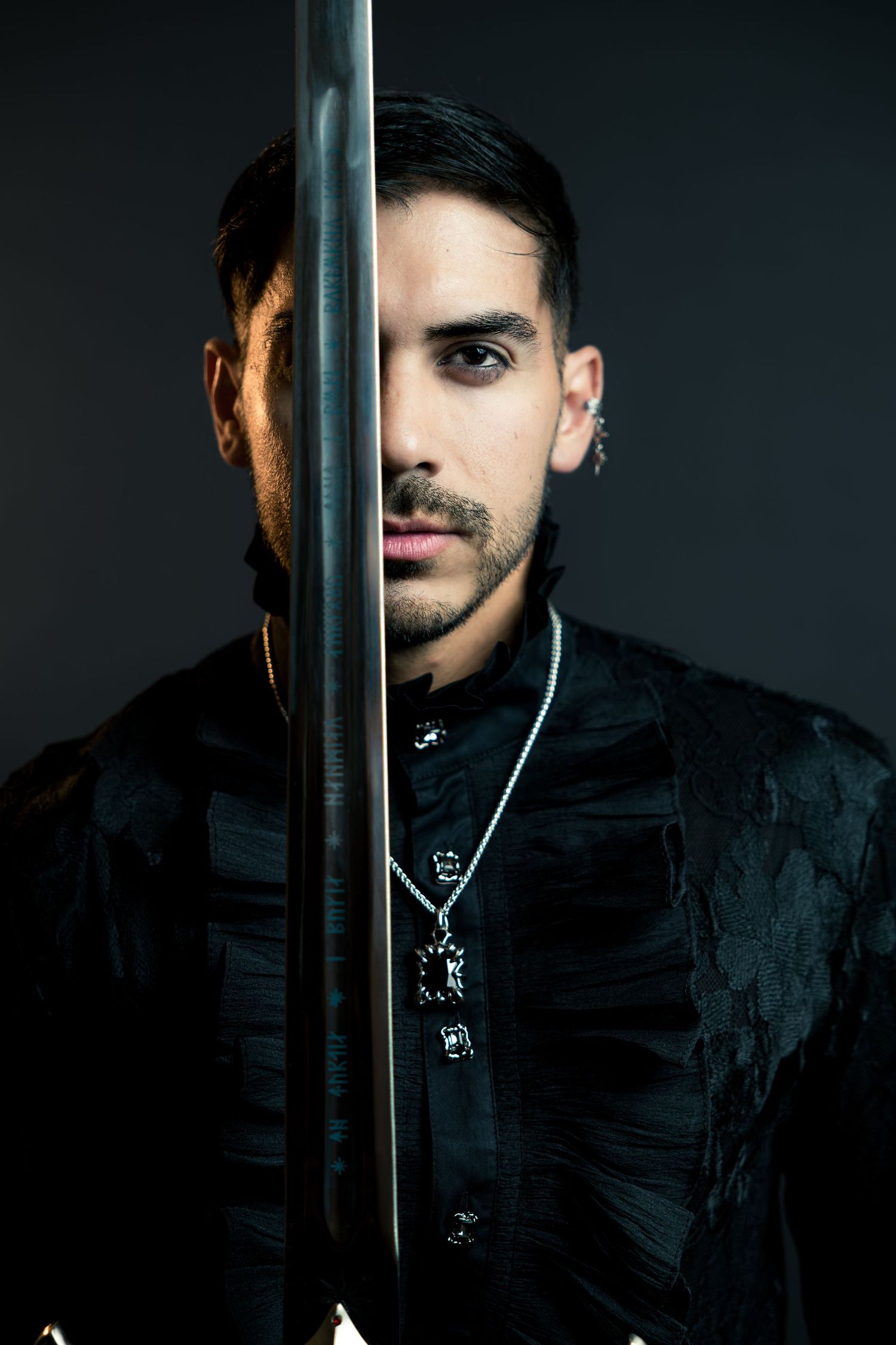 Man in black jacket holding sword vertically in front of face with chain necklace and ear piercing.