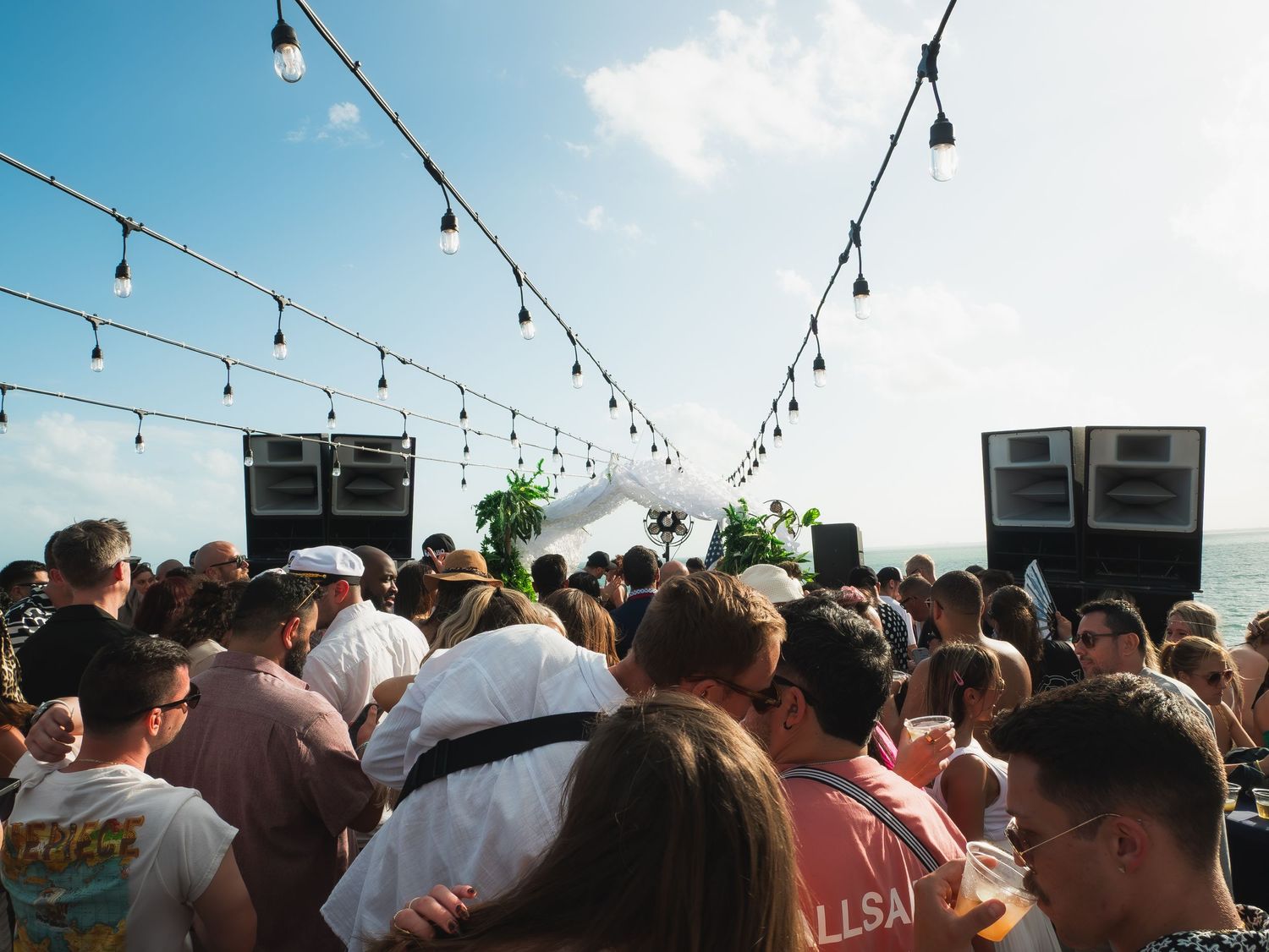 A crowded outdoor party scene with string lights overhead and large speakers set up during daytime.