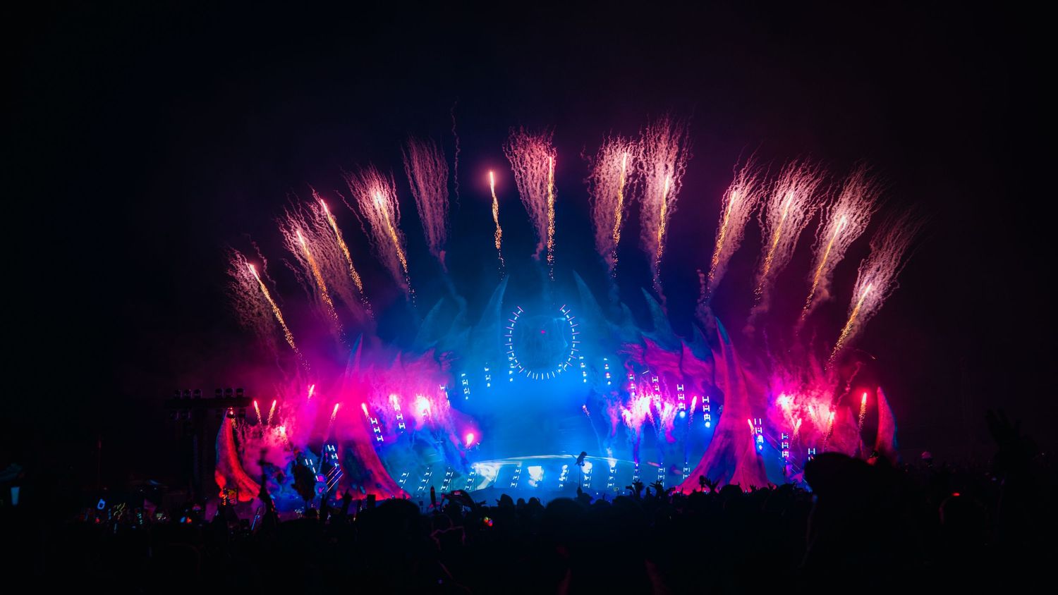 Spectacular fireworks display illuminates concert stage with vibrant pink and blue lights during nighttime performance.