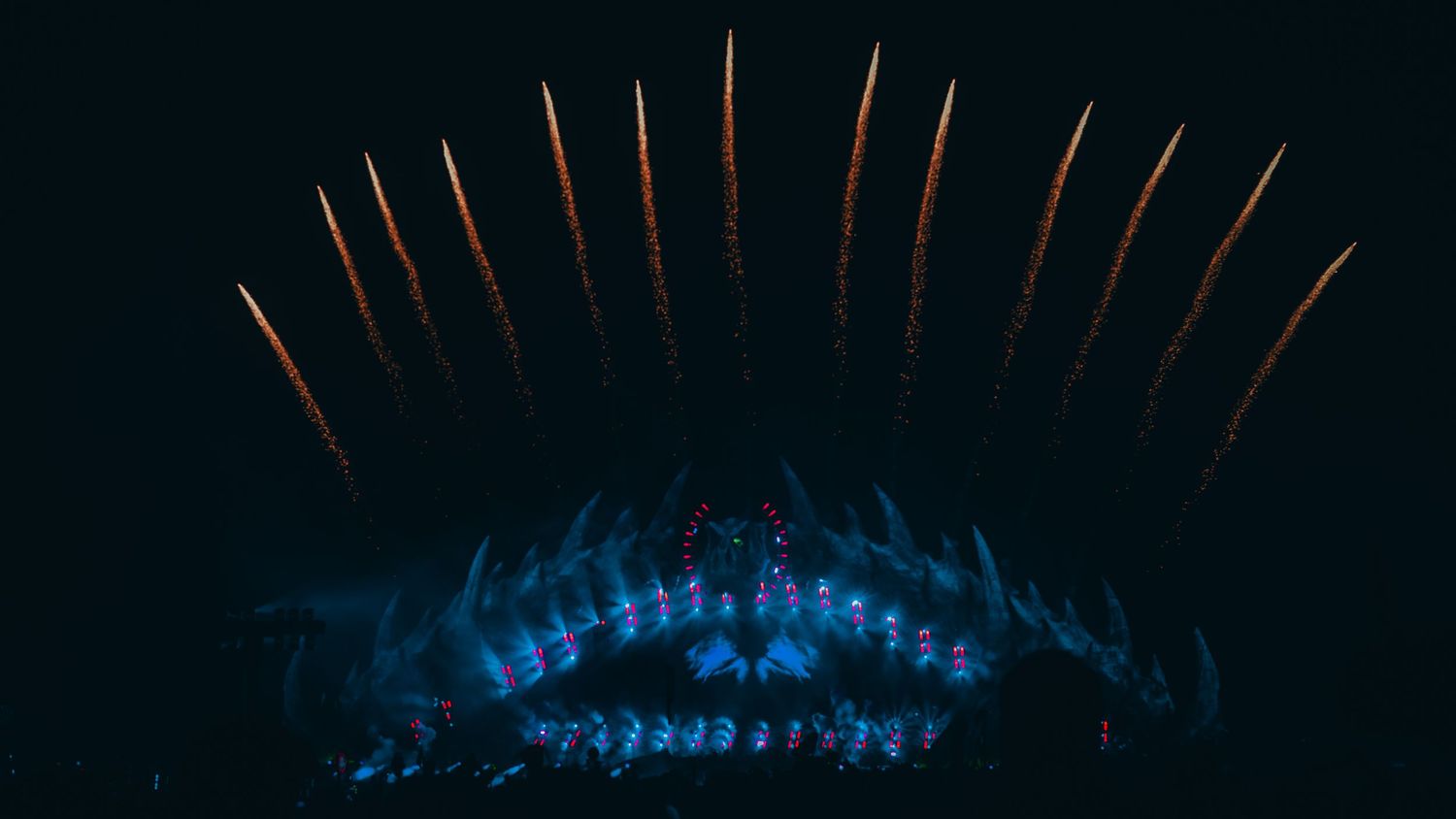 Blue-lit concert stage with golden pyrotechnic fountains shooting upward against a dark night sky.