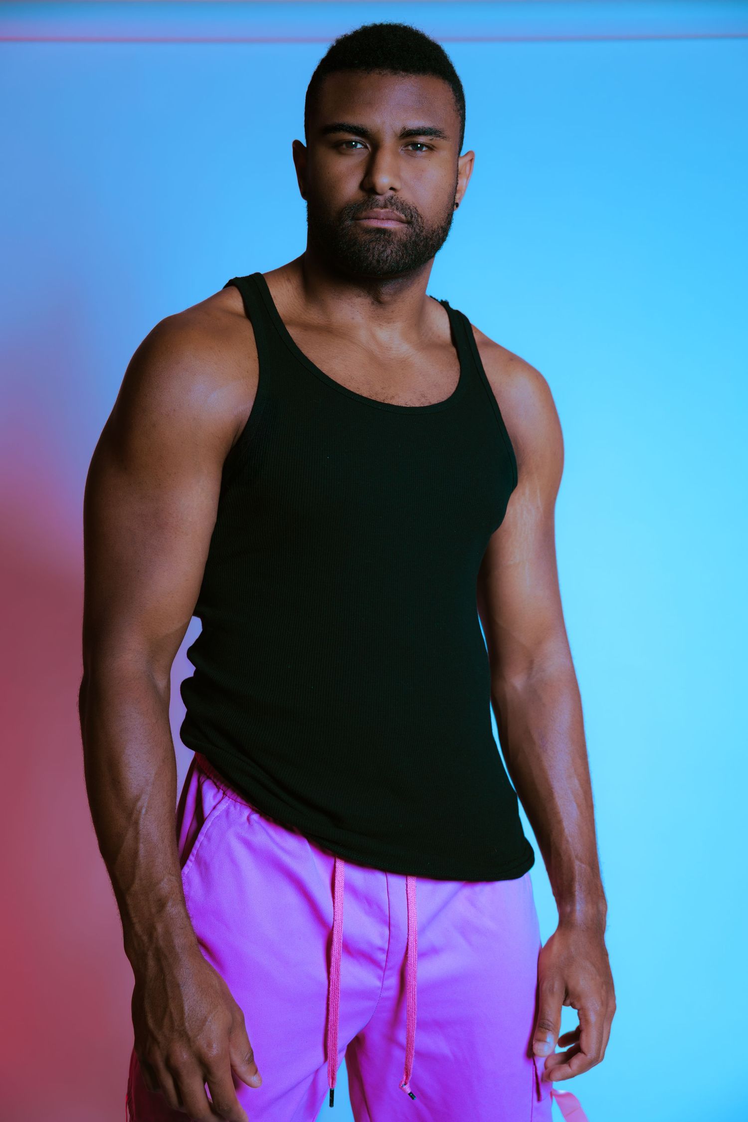 Muscular man in black tank top and pink shorts posing against blue and red neon background.