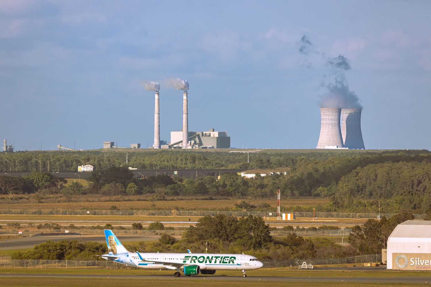 Frontier Airlines aircraft on runway with industrial power plant cooling towers in background.