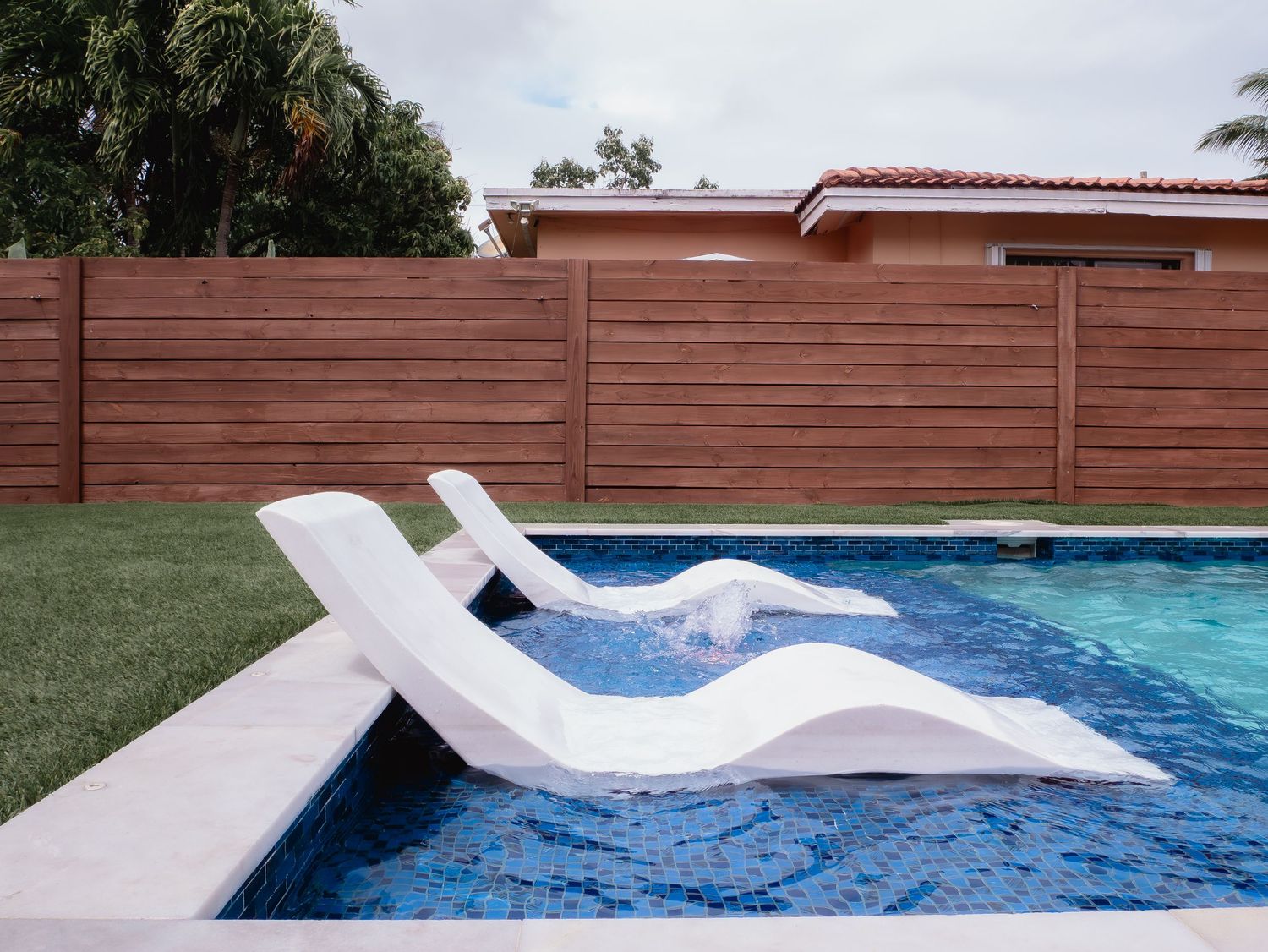White curved pool lounger partially submerged in swimming pool with wooden fence and artificial grass in backyard.