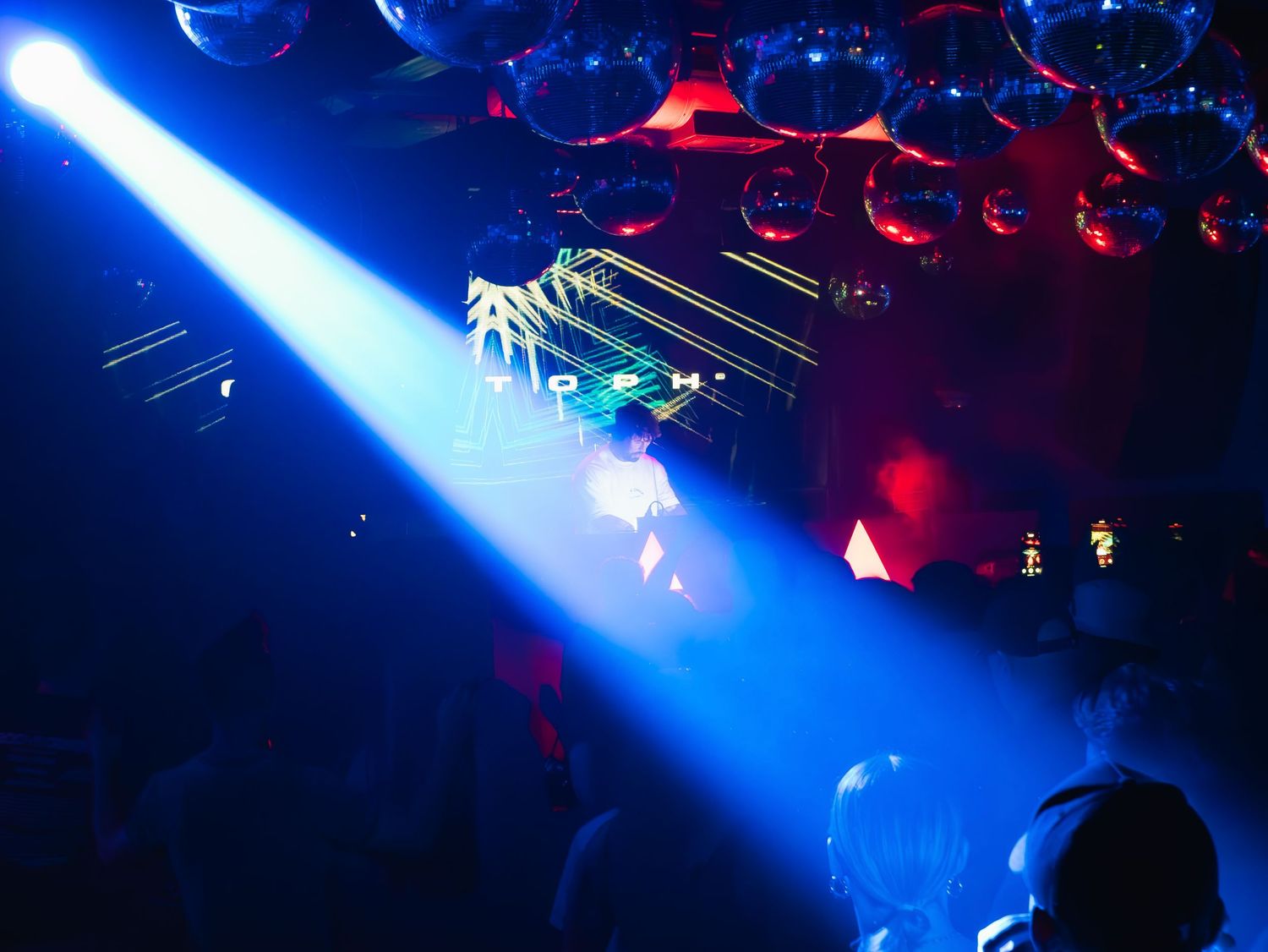 A bright blue spotlight beam illuminates a nightclub dance floor with red disco lights in the background.