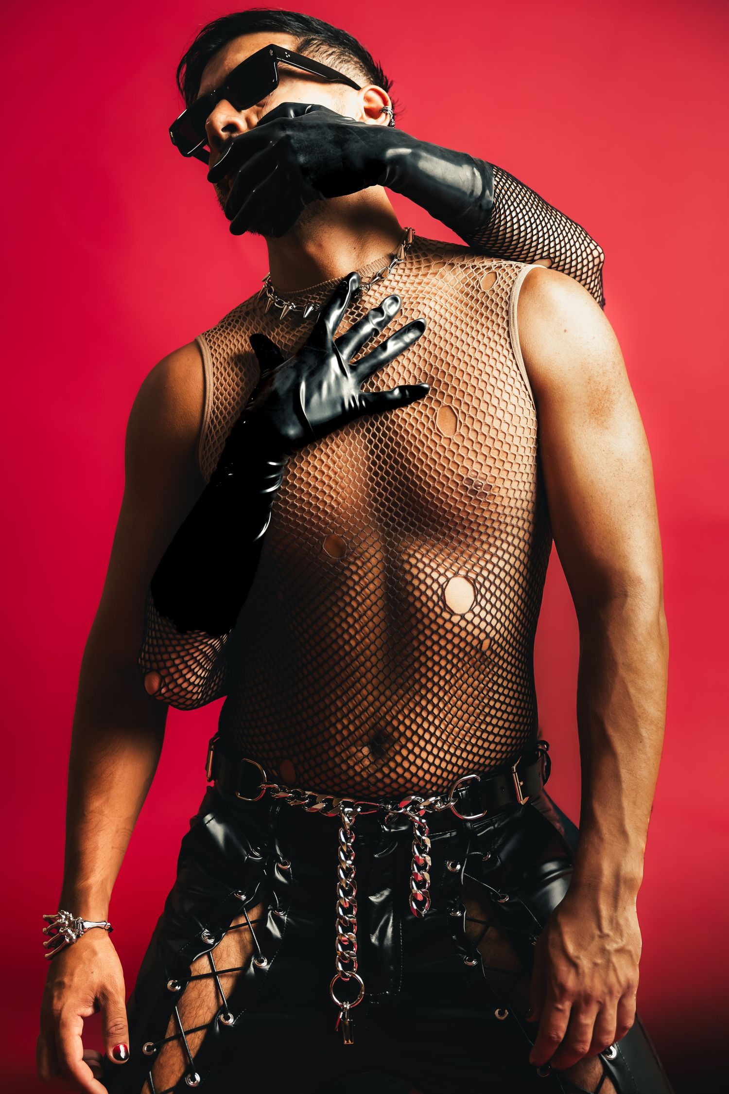 Muscular man in mesh top with black leather gloves and chain belt on red background.