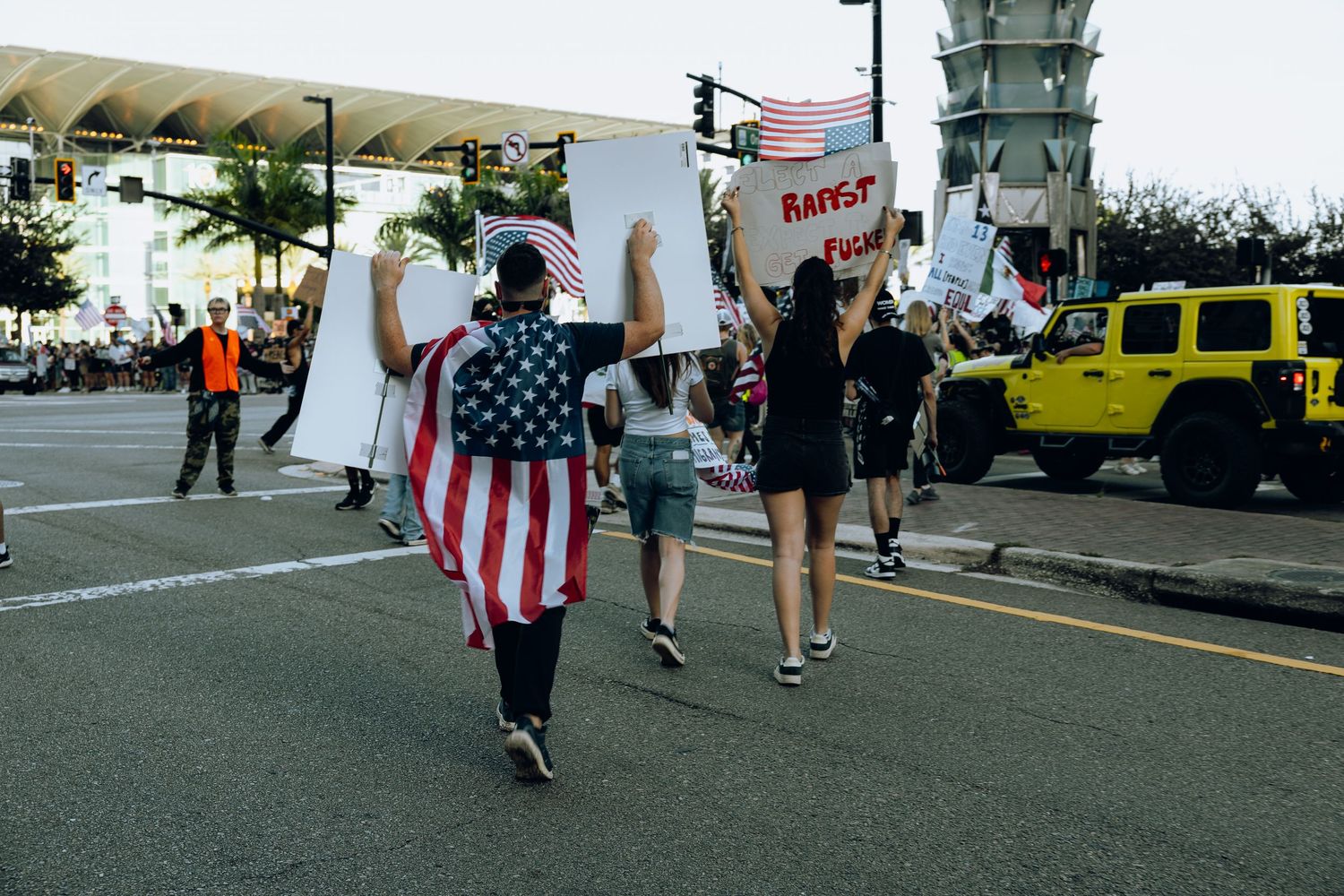 Person wearing American flag walks during protest march with yellow vehicle in background.