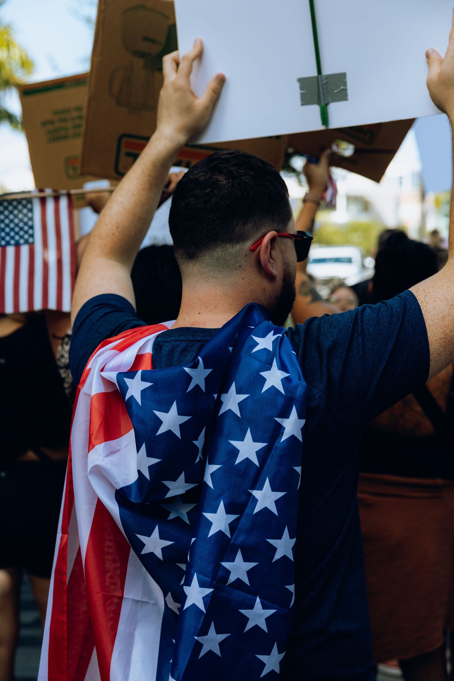 Protester wearing American flag attire raises arms at demonstration with US flag in background.