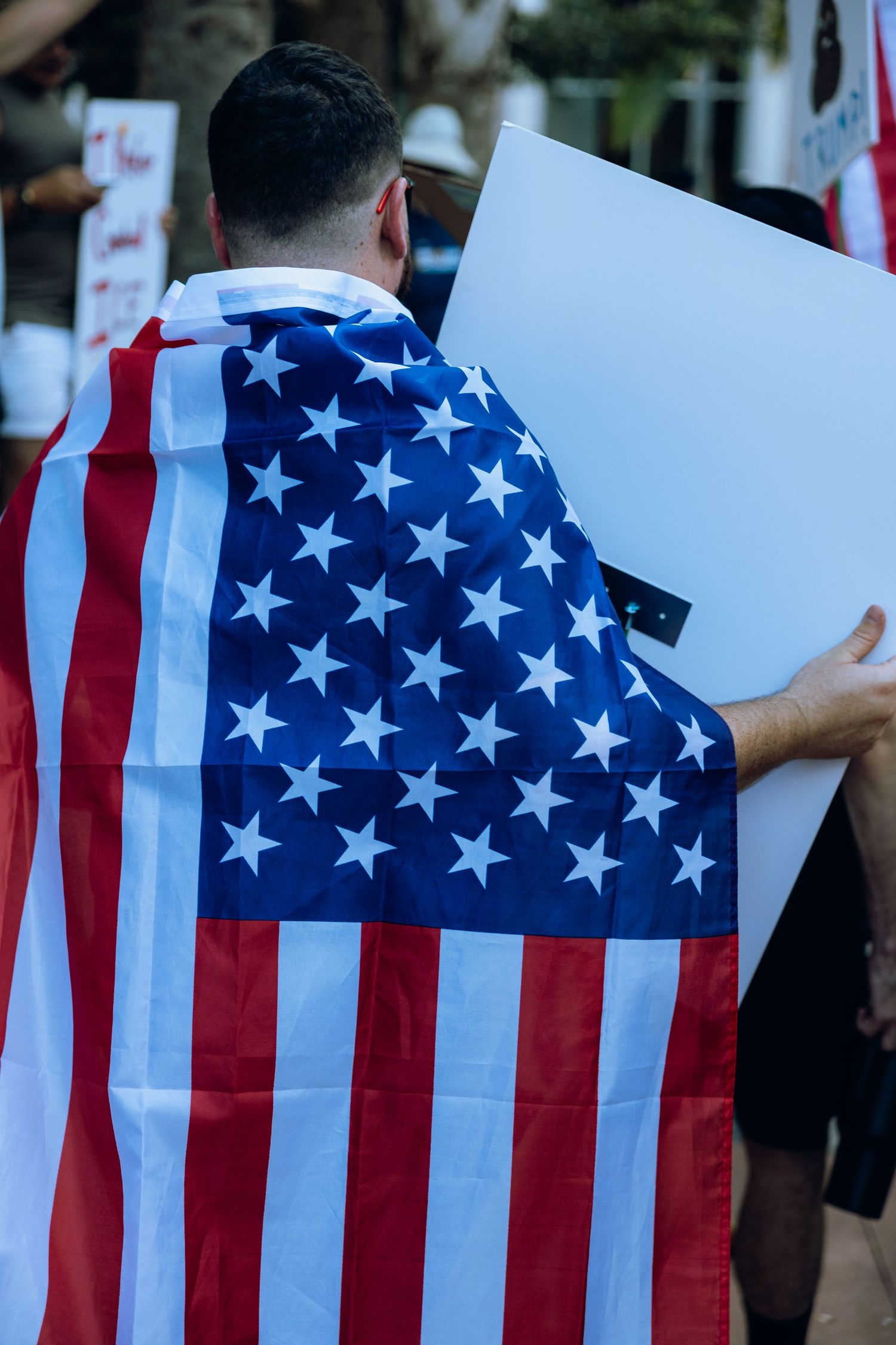 Back view of person wearing American flag as cape while holding white poster at rally.