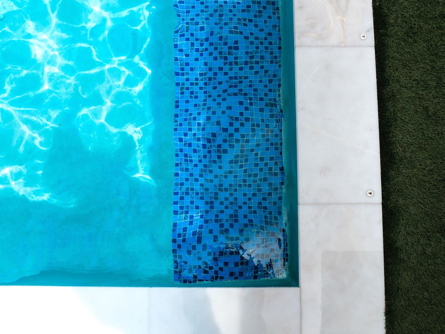 Aerial view of a swimming pool edge with blue mosaic tiles, white concrete border, and green grass.