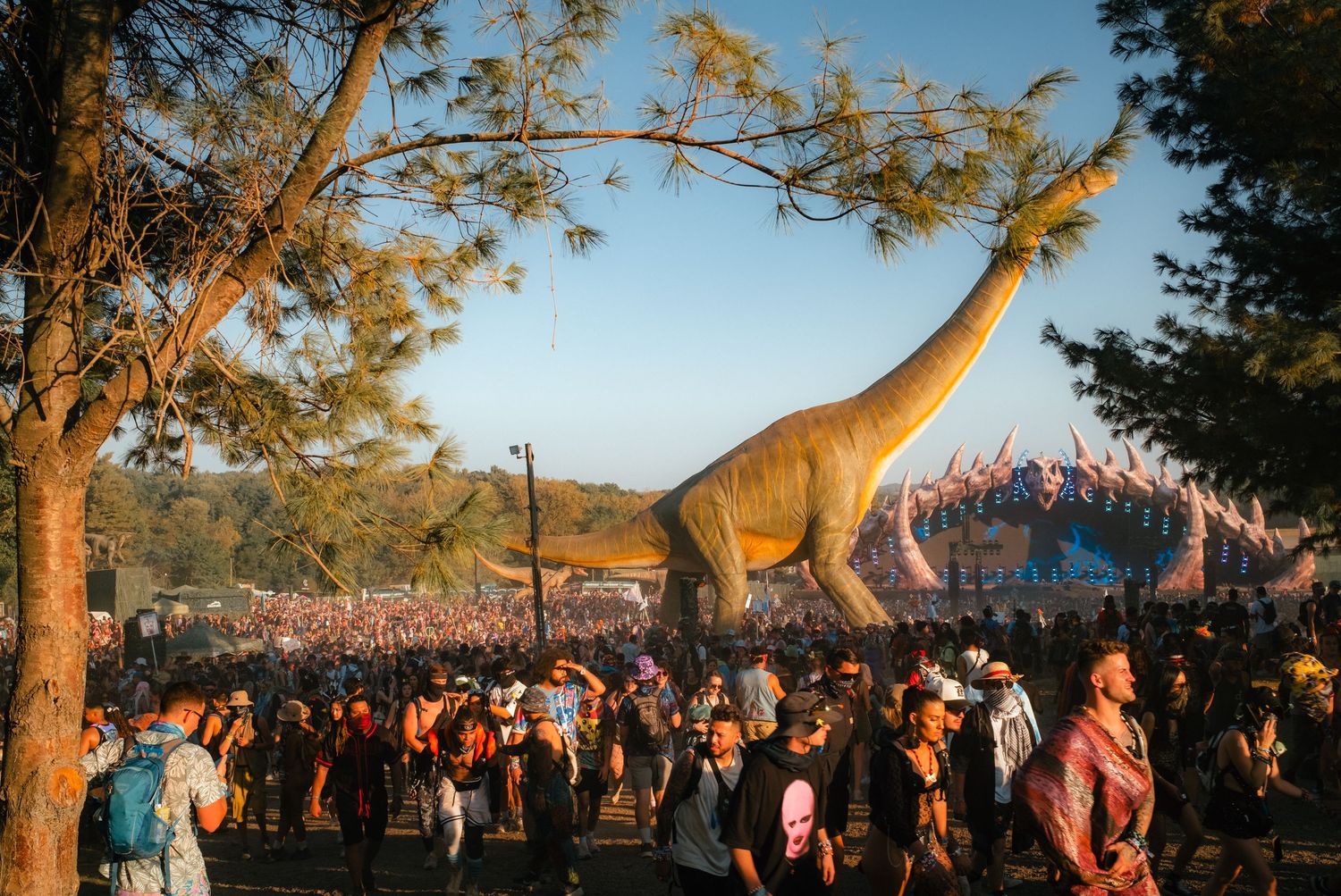 Large crowd gathers around a towering dinosaur statue at an outdoor festival during golden hour.