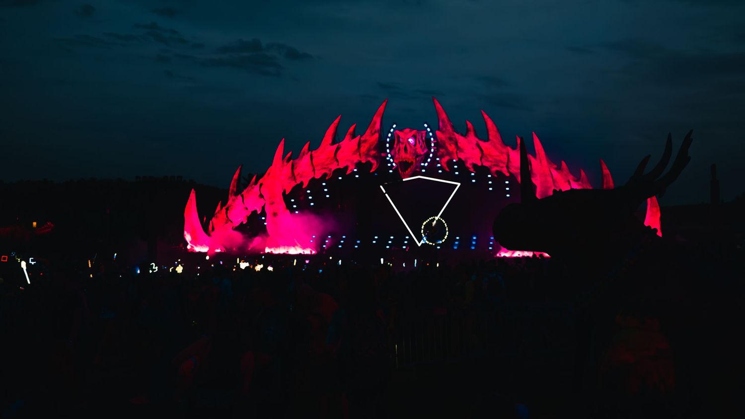 Pink neon lights illuminate a dark outdoor stage setup with geometric shapes and flame-like lighting effects.